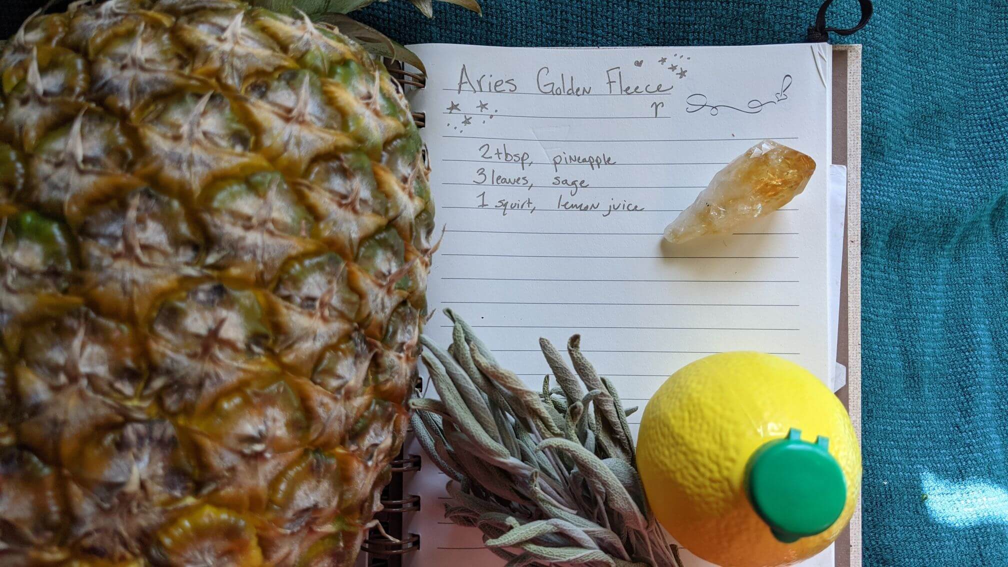 A fresh pineapple lays over a handwritten recipe. Dried sage, lemon juice, and a polished citrine quartz are featured.