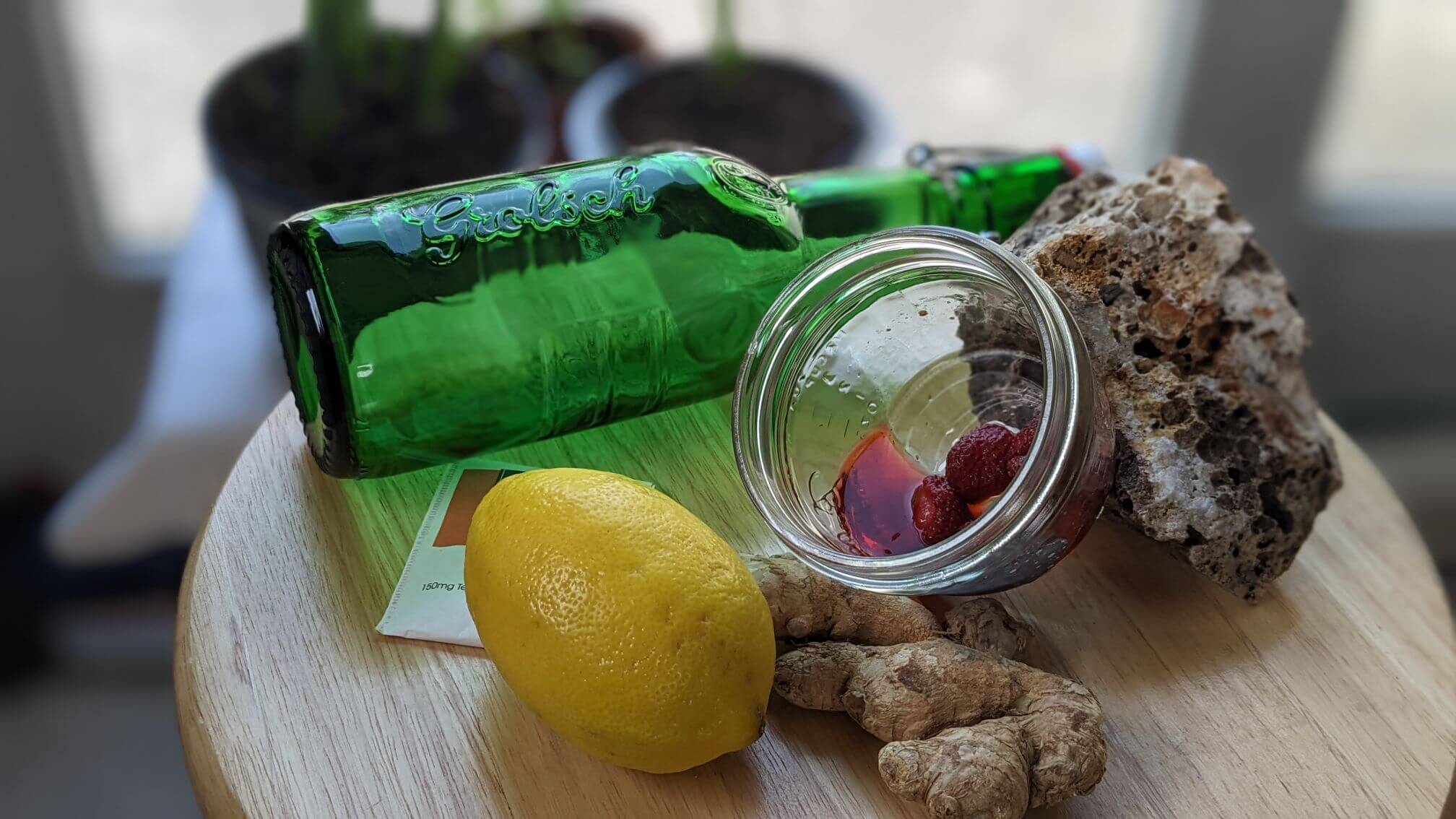 A fresh lemon sits next to thawed strawberries in a shallow dish. A green grolsch bottle sits in the background with a fresh ginger root and a natural stone with small fossilized areas.