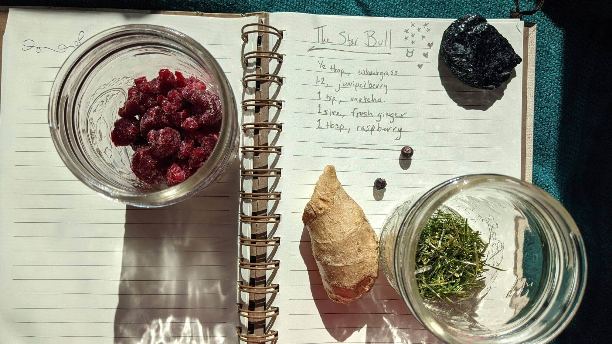 Handwritten recipe shown in bright daylight. Thawing frozen raspberries in a jar, a scattering of juniper berries, fresh ginger, and dehydrated wheatgrass are featured.