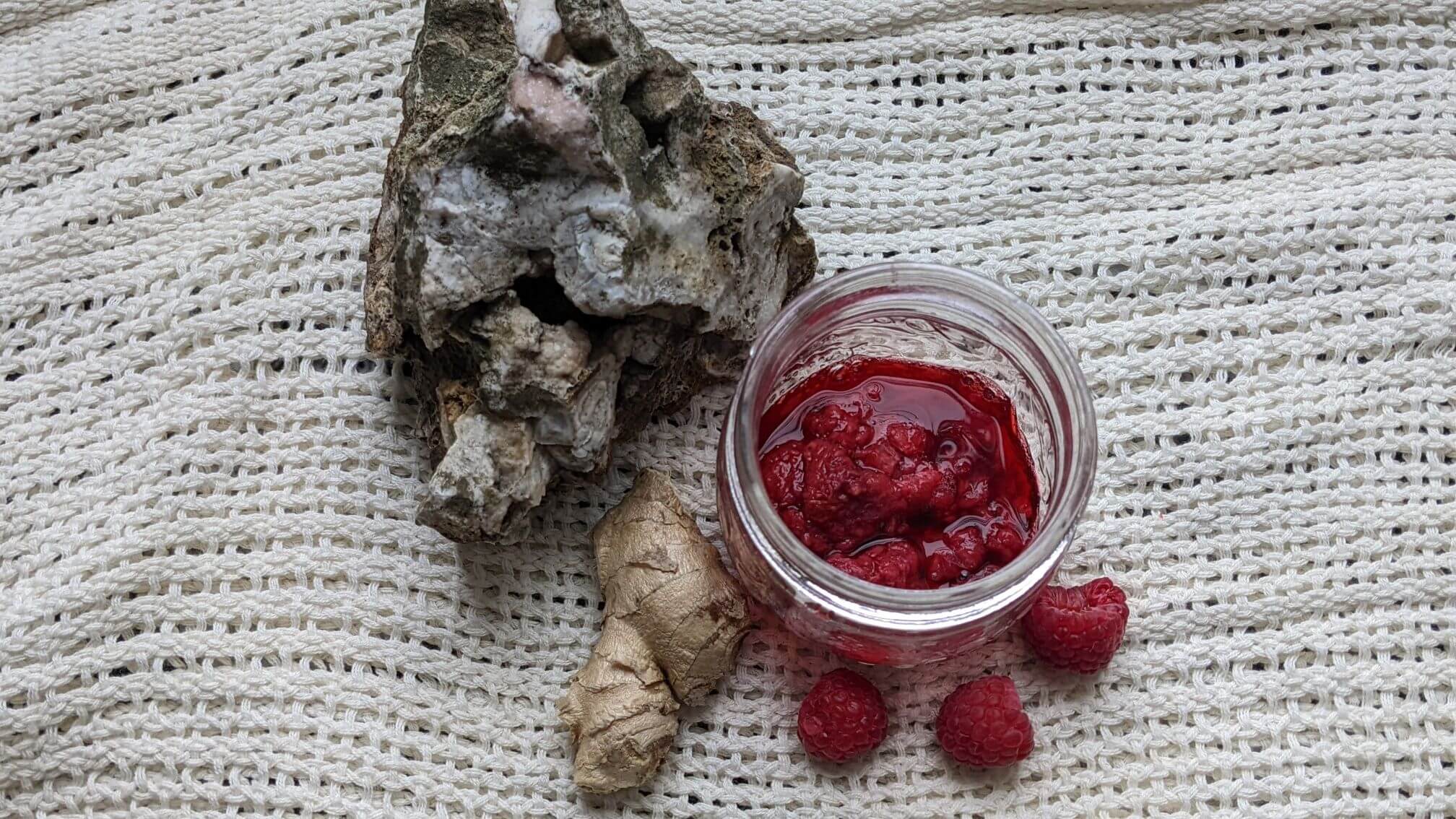 A jar of thawed, frozen raspberries sits on a linen scarf with fresh raspberries scattered around a fresh ginger root. A natural rock with druzy quartz vein flanks the image.