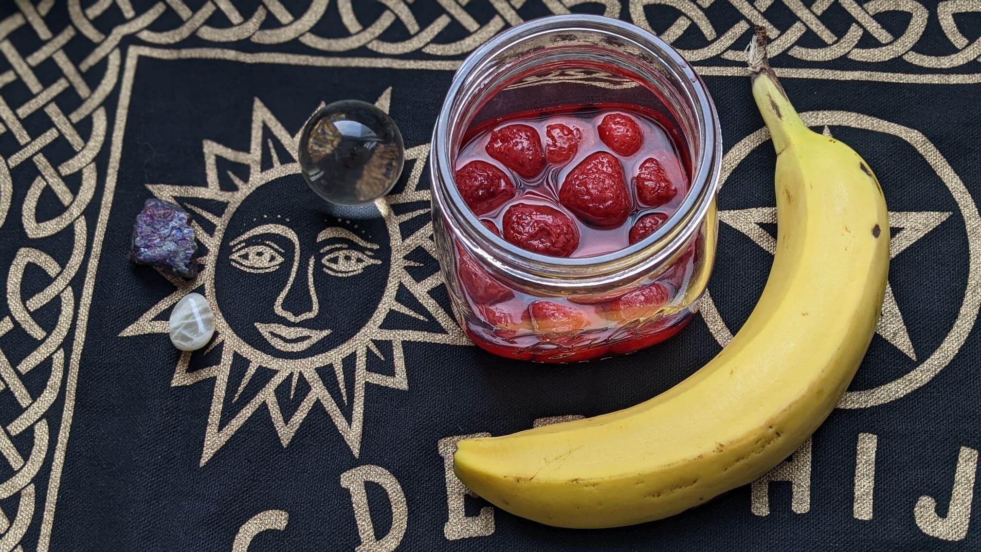 Strawberries sit in their own juice in a glass jar on a table with a Ouija Board cloth. A full, fresh banana and a few gems are also on the cloth.