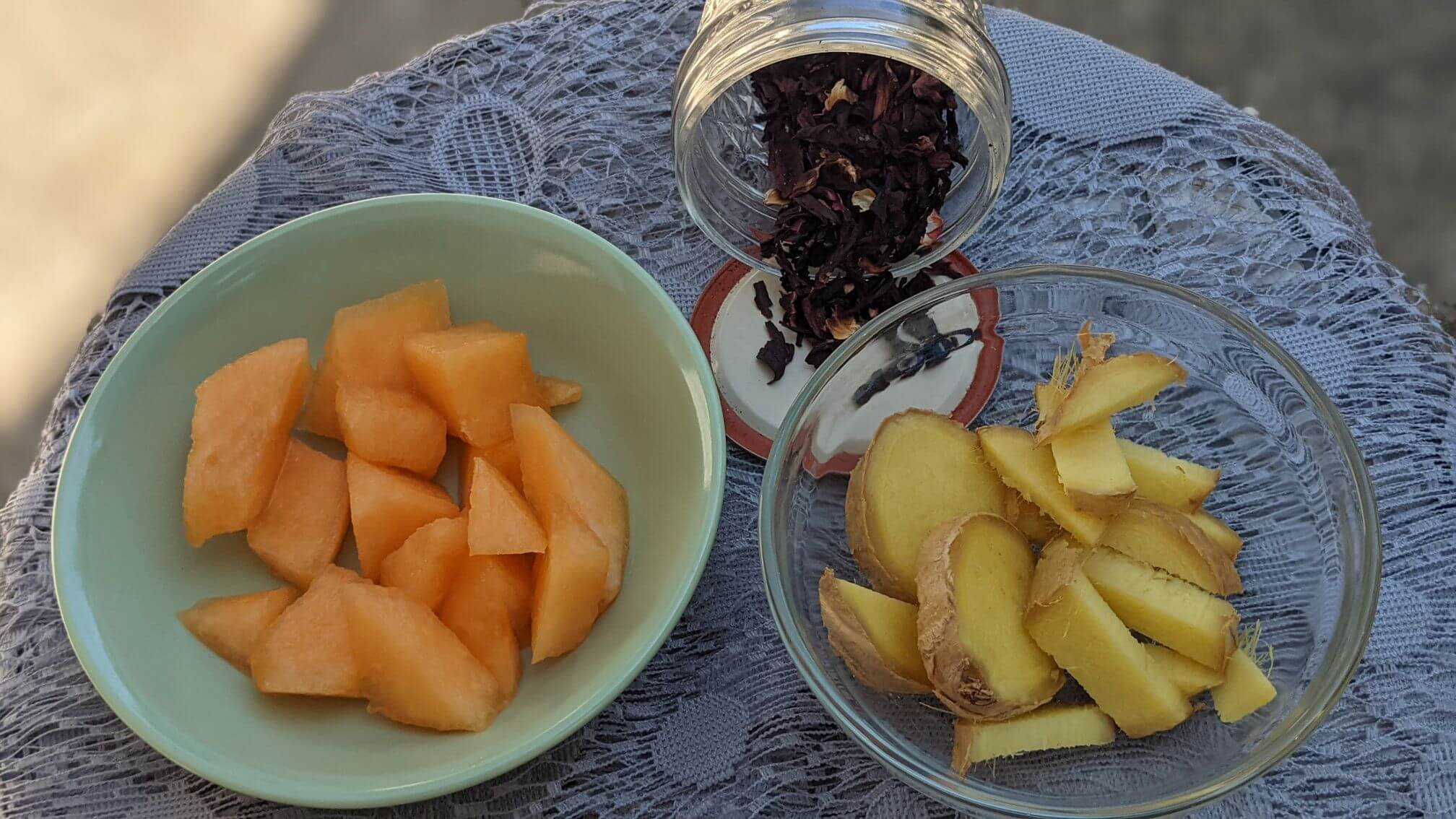 Fresh melon is in a shallow dish next to a scattering of dried hibiscus petals and sliced fresh ginger.