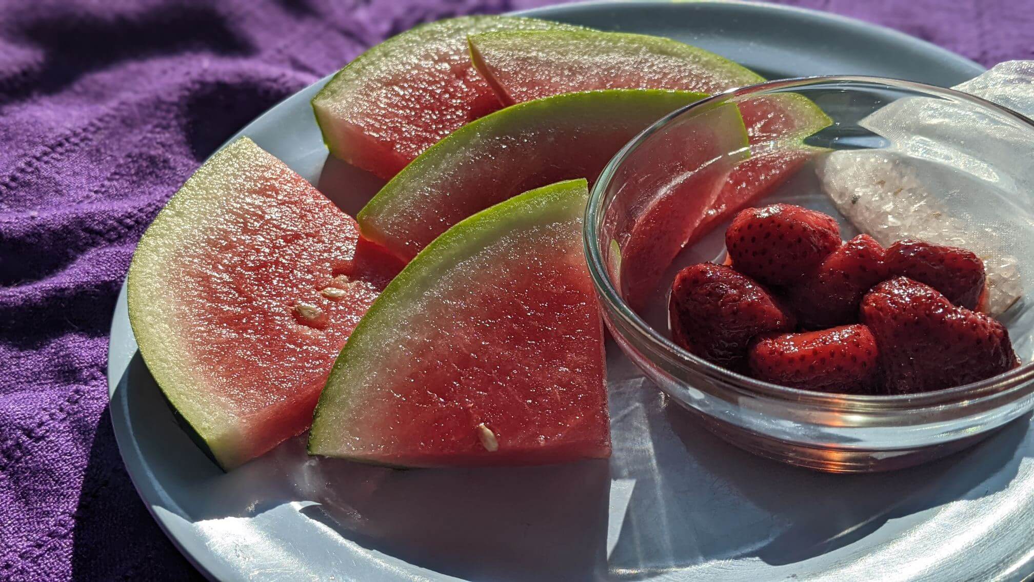 Fresh watermelon sit on a plate in the sun, with a shallow dish of thawed frozen strawberries.