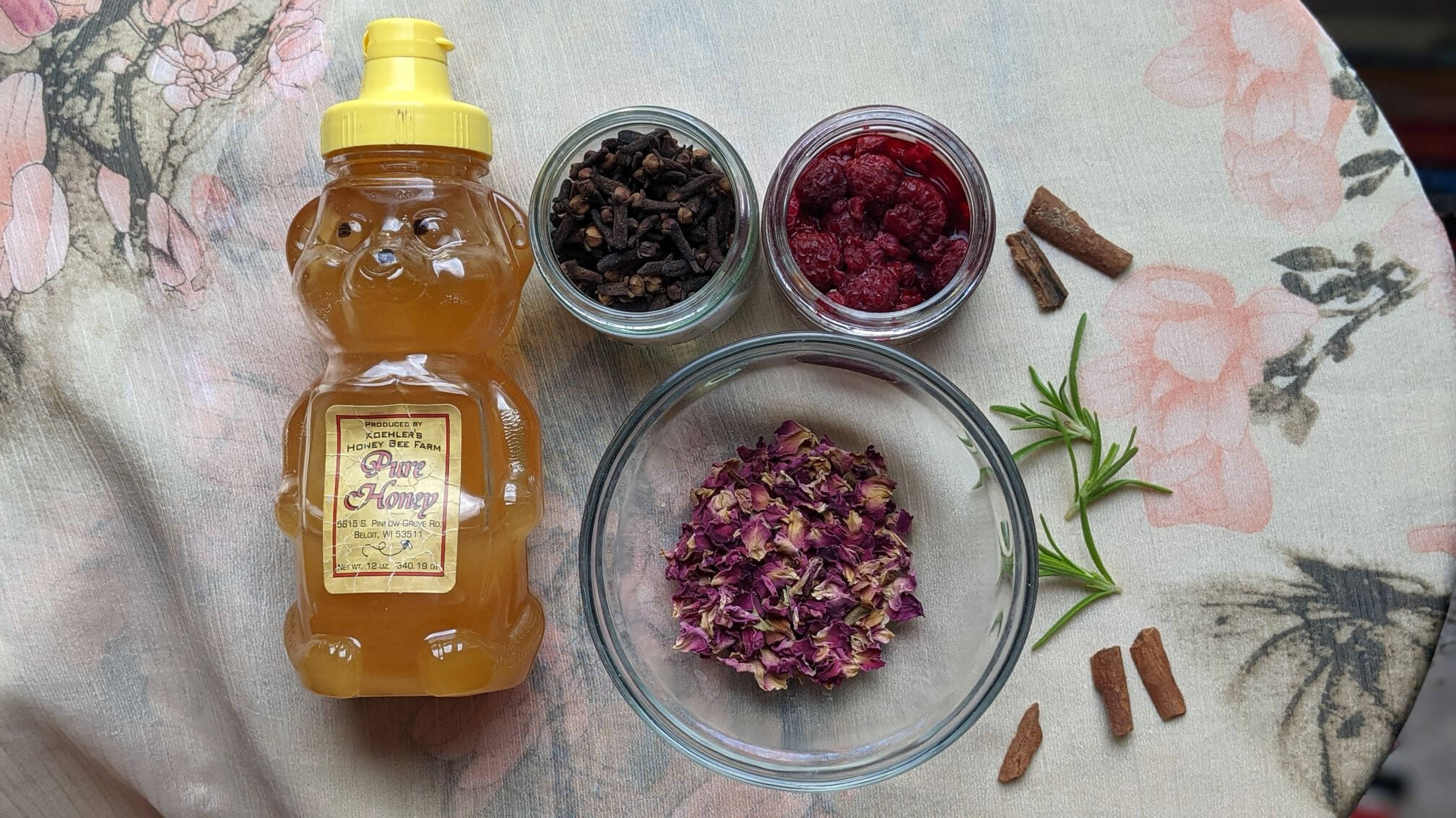 In natural light, a bear-shaped bottle of honey lays on a table next to shallow bowls containing dried rose petals, thawed frozen raspberries and cloves. Scattered next to the bowls are small stalks of cinnamon and a few sprigs of fresh rosemary.