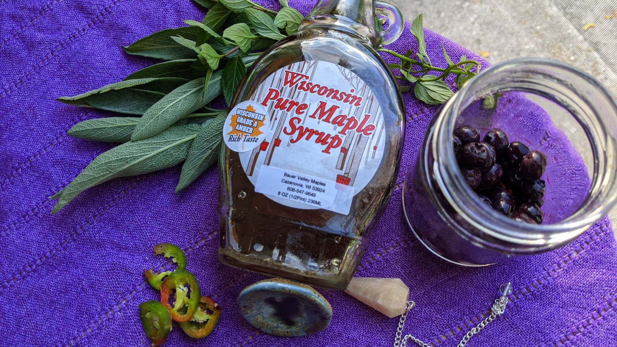 A small jar of thawed, frozen blueberries sits on a purple tablecloth with a jar of Wisconsin maple syrup, a few slices of fresh jalapeno pepper, and a moonstone pendant.