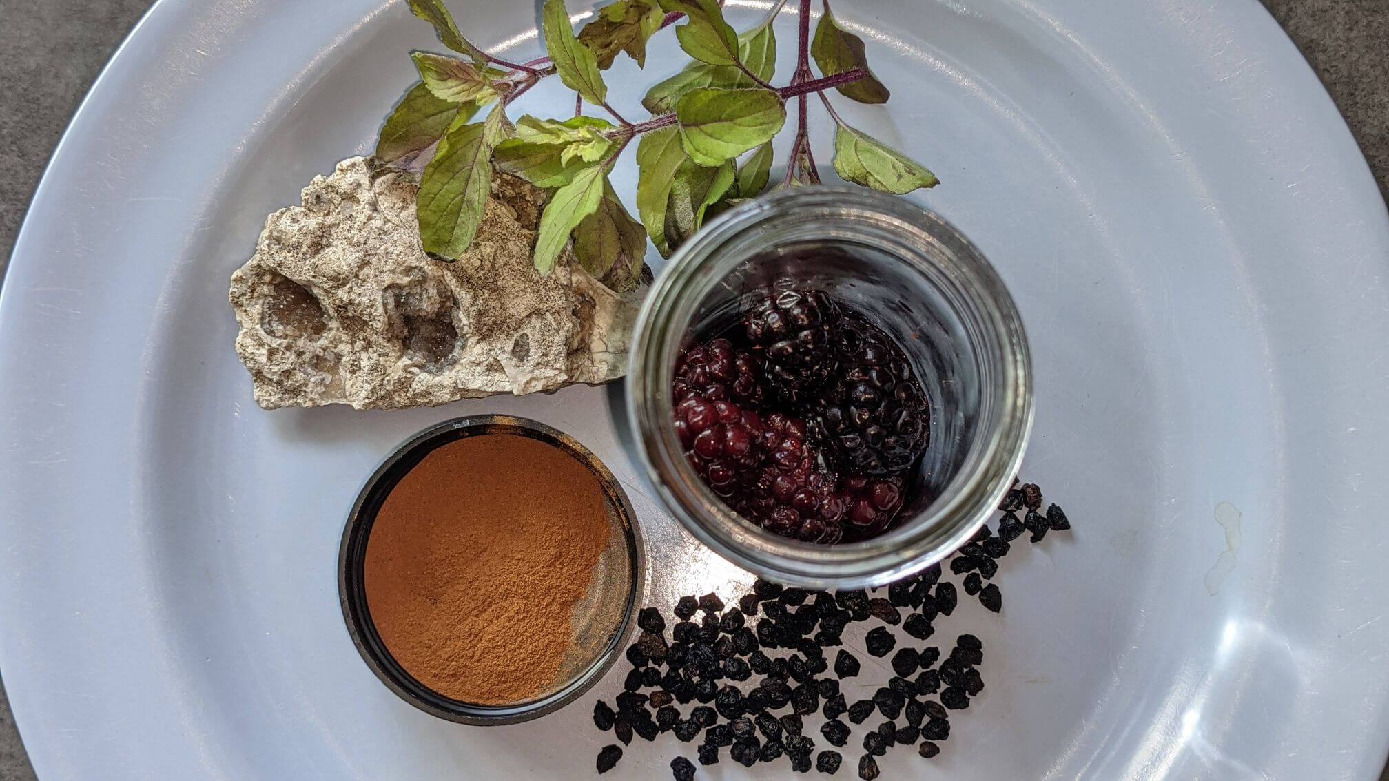 Thawed, frozen blackberries in a glass jar sit on a blue plate with a jar of cinnamon powder. Scattered next to the jars are dried elderberries. A druzy quartz and fresh tulsi are also on the plate.