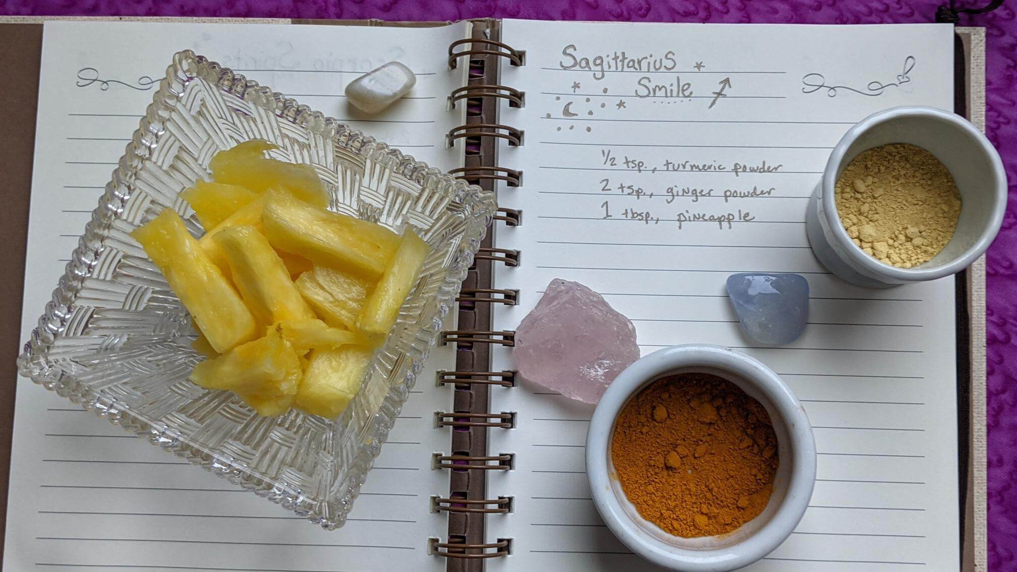 The handwritten recipe is featured with a crystal bowl filled with sliced pineapple. A rose and smoky quartz are scattered on the book with two shallow bowls of dried ginger and dried turmeric powder natural light.