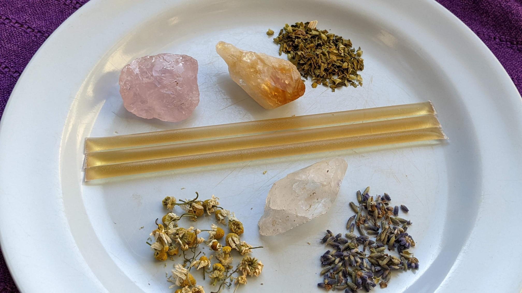 A rose quartz and citrine sit on a plate next to three honey straws. A small pile of dried chamomile, dried lavender, and dried skullcap adorn the plate in natural light.
