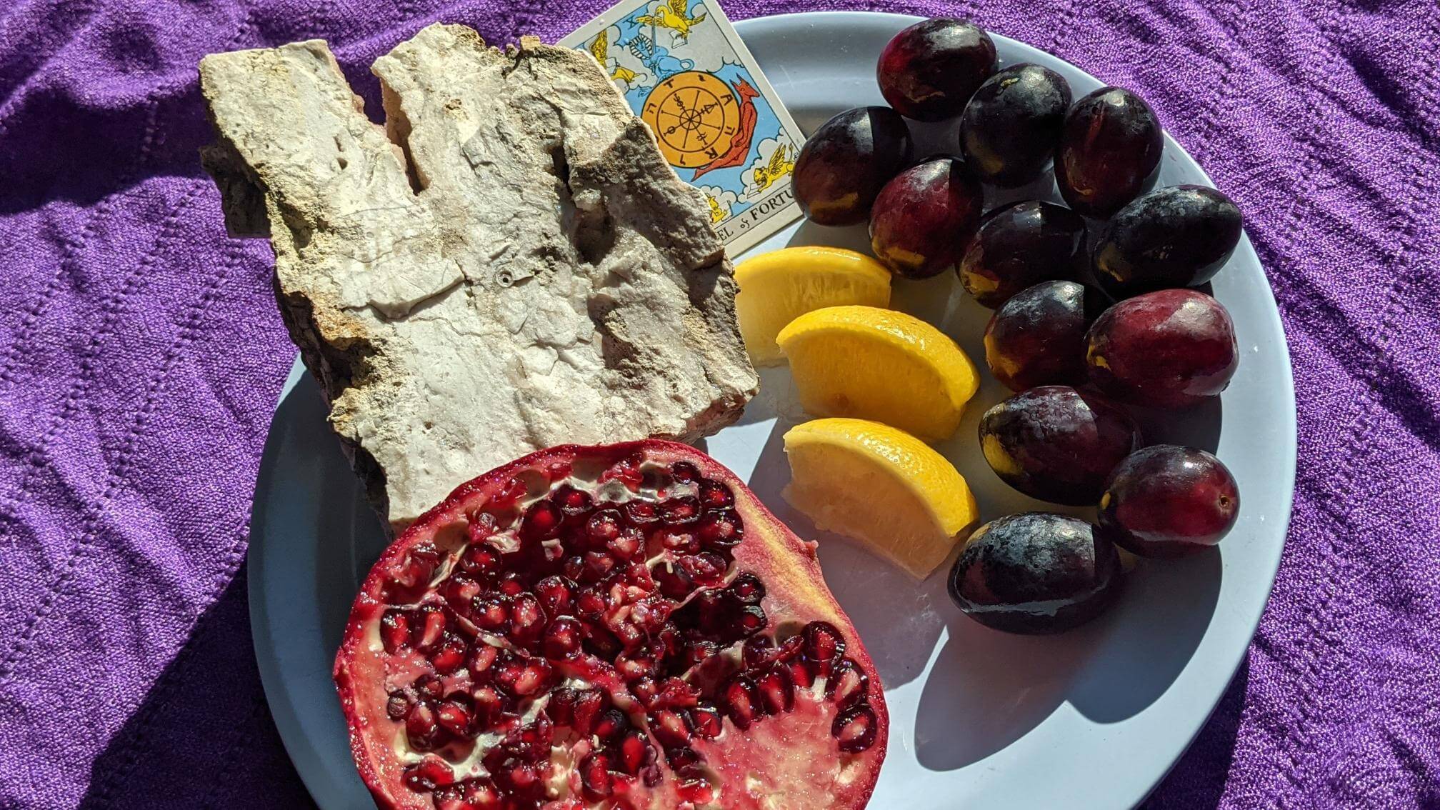 In natural daylight on a purple tablecloth, a fresh pomegranate is cracked open. Next to it on a blue plate, a chunk of rock with a quartzite notch, fresh slices of lemon and 12 red grapes sit by a jar of honey.